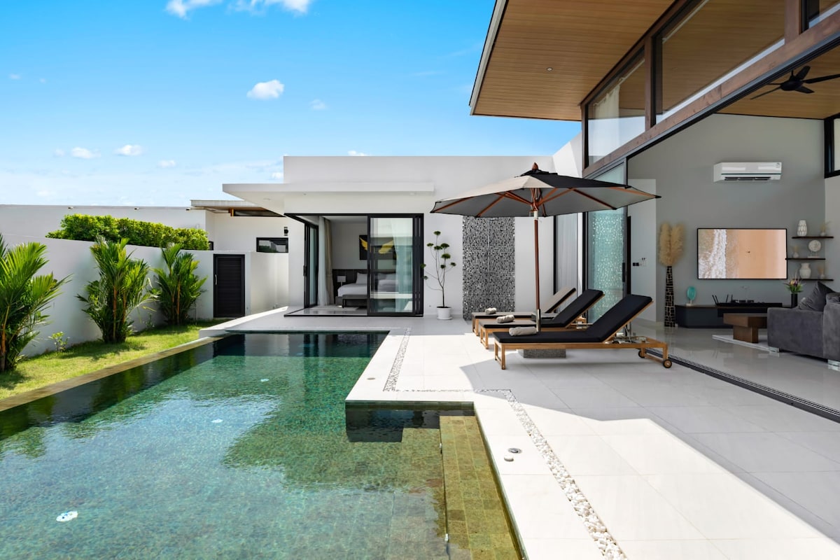 A contemporary villa exterior features a private pool and an outdoor seating area with lounge chairs under a large umbrella. Lush greenery surrounds the pool, and expansive glass doors provide views into the living space, inviting natural light into the home.