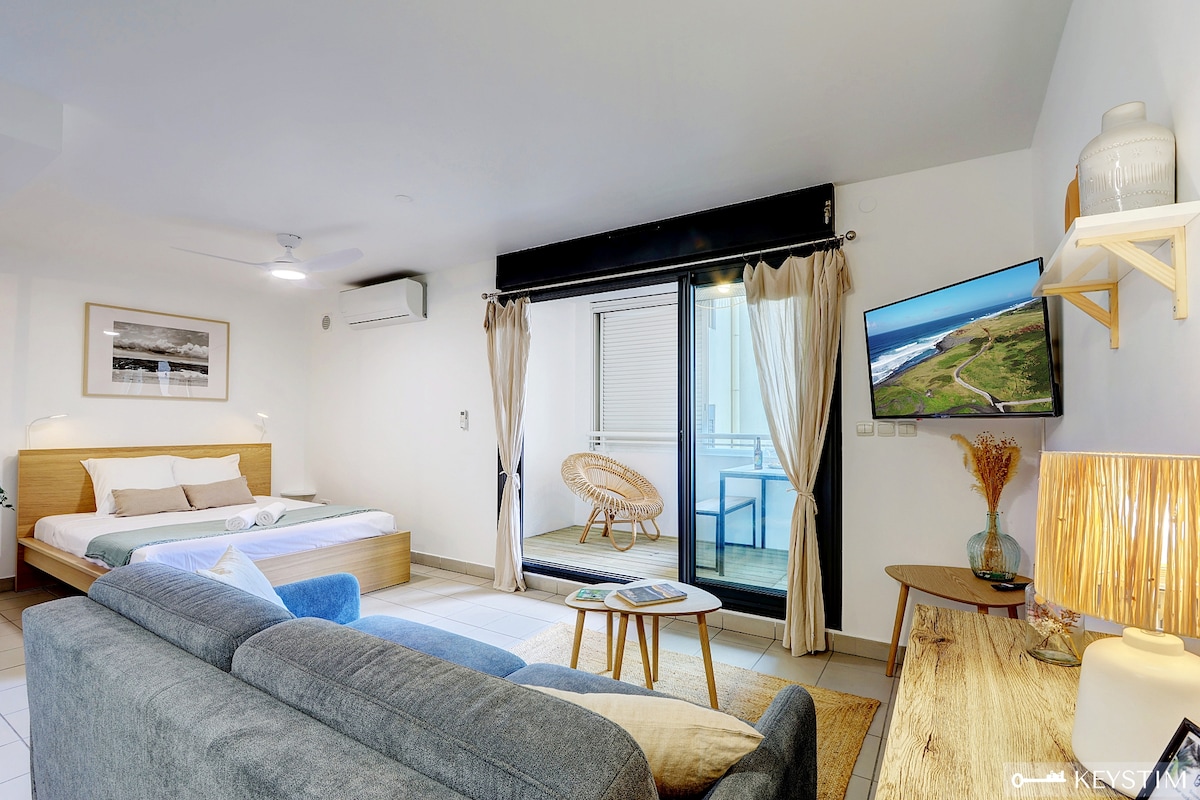 A modern studio space features a spacious bed with neutral bedding and a cozy sofa. A flat-screen TV is mounted on the wall opposite the bed. Large sliding glass doors open to a balcony area, adorned with light curtains allowing natural light to fill the room.