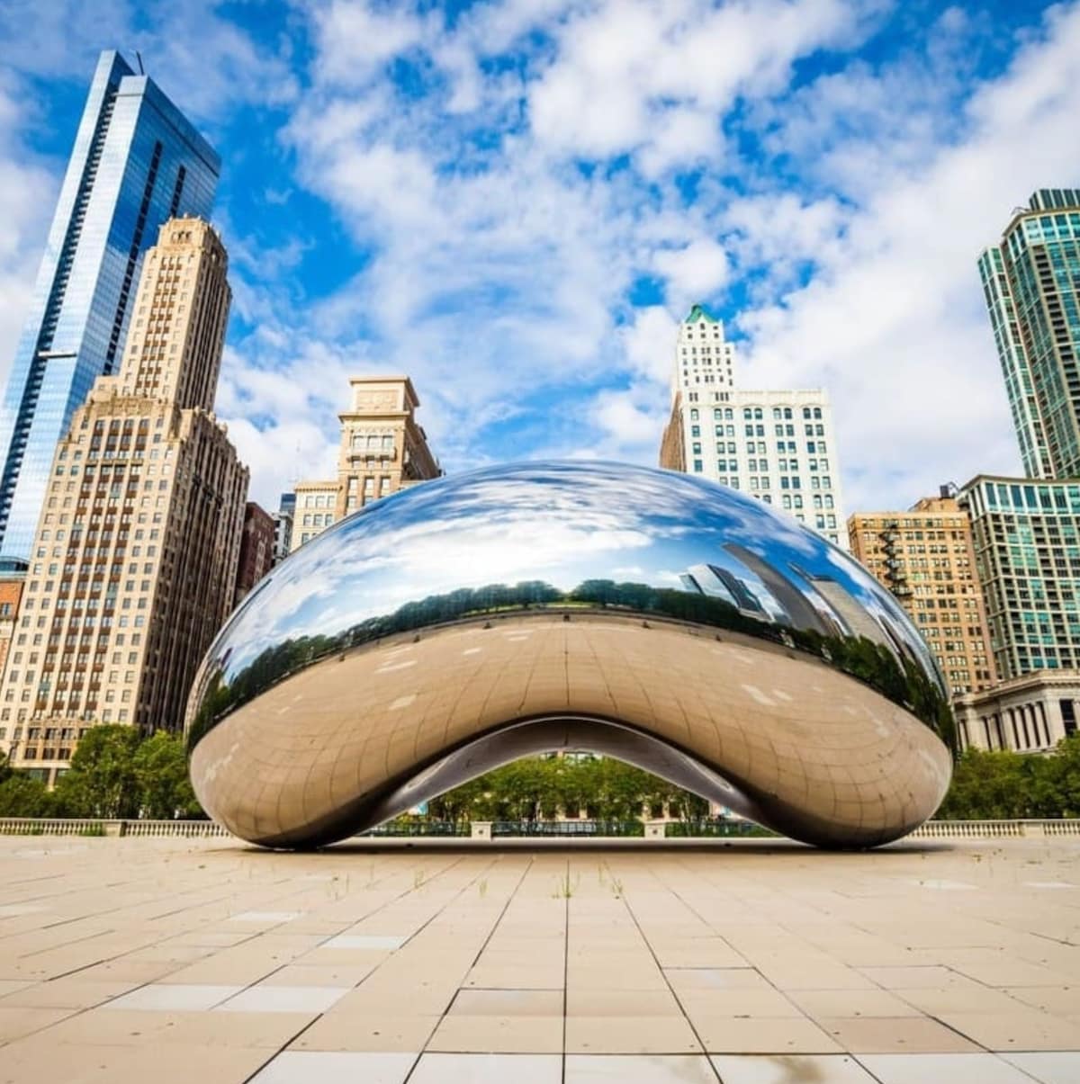 A large, reflective sculpture known as The Bean is shown in a public plaza. Surrounding skyscrapers and a bright sky with scattered clouds are mirrored on its surface, creating a dynamic urban scene. A paved area leads up to the sculpture, enhancing its prominent position.