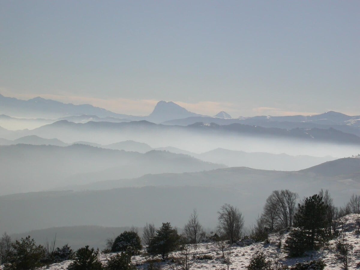 A panoramic view of misty mountains is displayed, showcasing multiple layers of slopes fading into the distance. The snowy peaks are partially obscured by a gentle haze, offering a serene and tranquil landscape. Sparse trees and shrubs can be seen in the foreground.