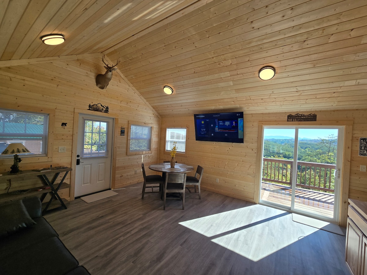A warm wood-paneled living area features a central dining table next to a large glass door leading to a deck. A flat-screen TV is mounted on a wall. Natural light pours in through multiple windows, enhancing the spacious feel of the room.