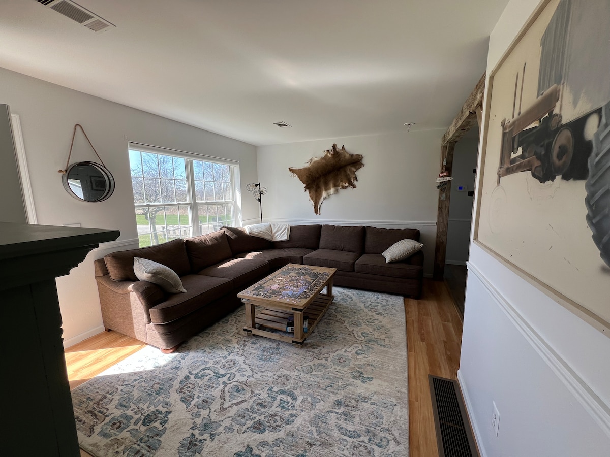 A spacious living area features a large brown sectional sofa, complemented by a wooden coffee table set on a patterned rug. Natural light enters through a window adorned with light curtains, illuminating the room. A circular mirror and a wall art piece enhance the decor.