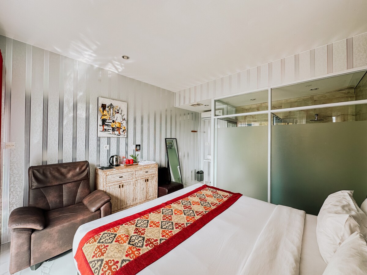 A comfortable room features a spacious bed with a decorative blanket, accompanied by a seating area with a brown armchair. A wooden dresser with a mirror is present, along with a kettle for refreshments. The bathroom is separated by frosted glass partitions, enhancing the room's openness.