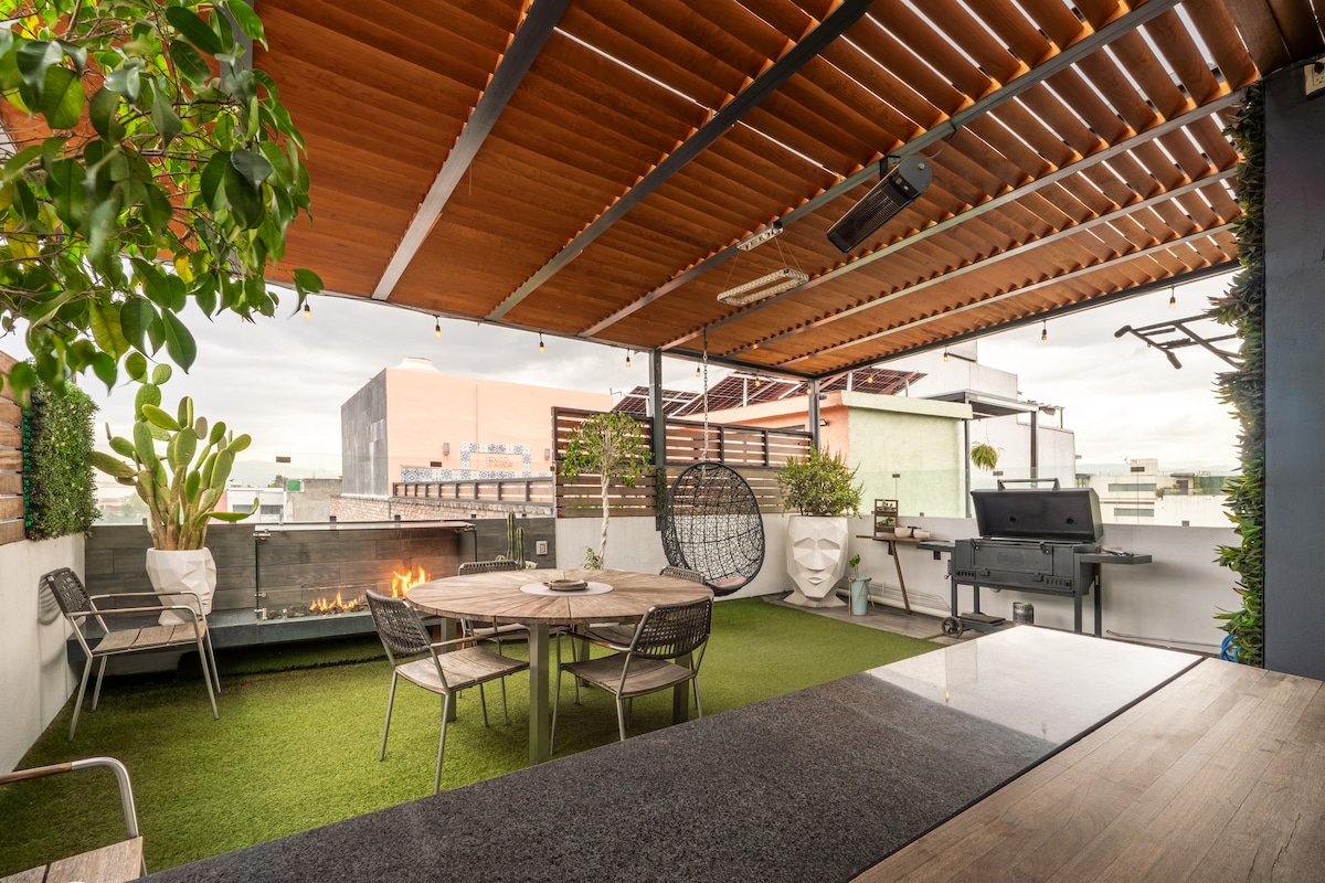 A private rooftop terrace features a wooden pergola overhead, providing shade and ambiance. A circular dining table with several chairs is centered on the space, alongside a grill and an automatic fireplace. Lush greenery enhances the atmosphere, complemented by a cozy hanging chair.