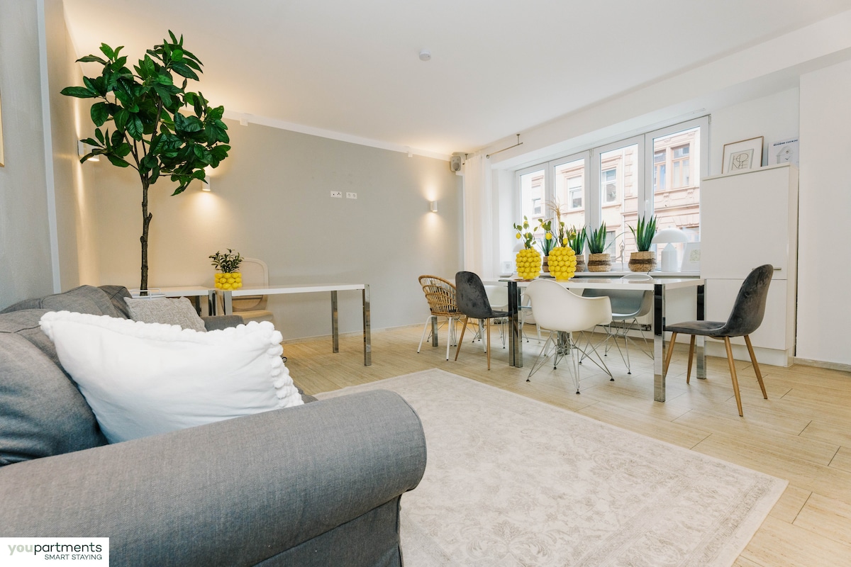 A modern living area is filled with natural light from large windows. A light rug adorns the floor, complemented by a gray sofa and a sleek dining table surrounded by colorful chairs. Decorative plants and vibrant yellow centerpieces add a touch of freshness to the space.