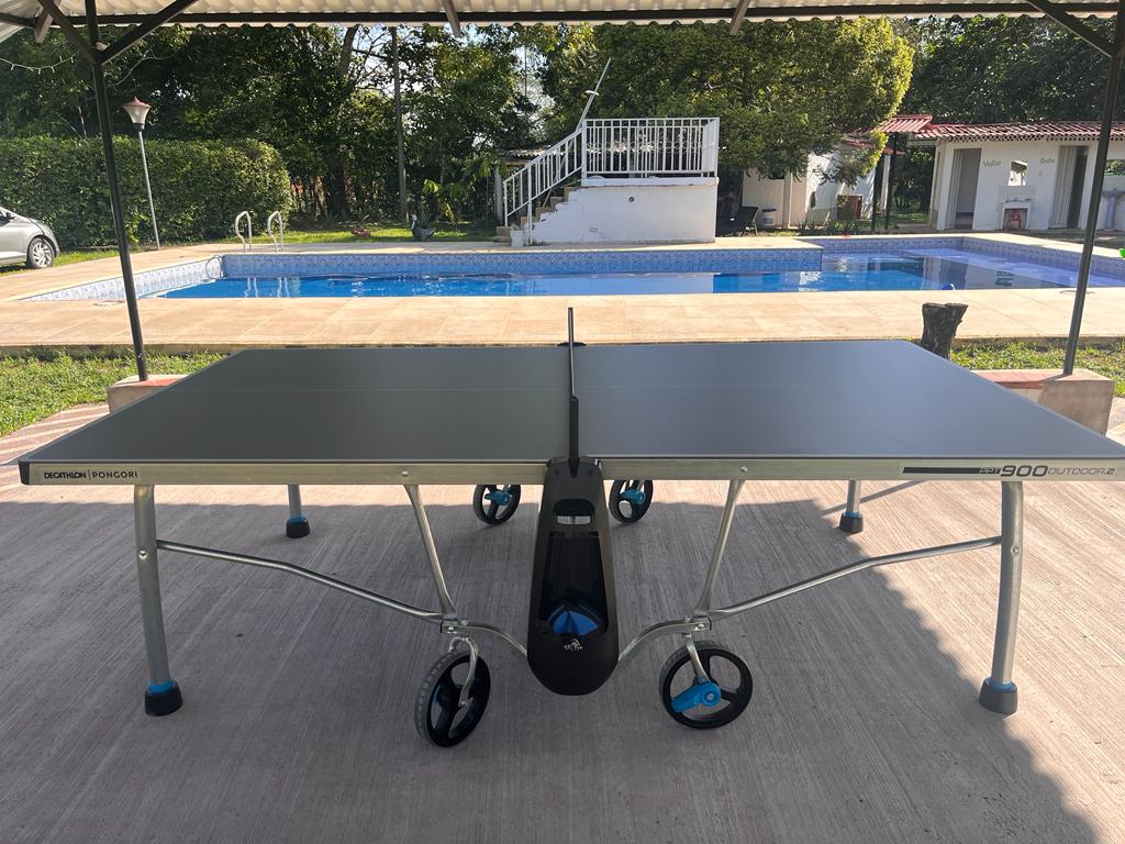 A modern table tennis table is positioned under a shaded area, ready for play. A sparkling swimming pool and green outdoor space are visible in the background, adding to the recreational atmosphere.