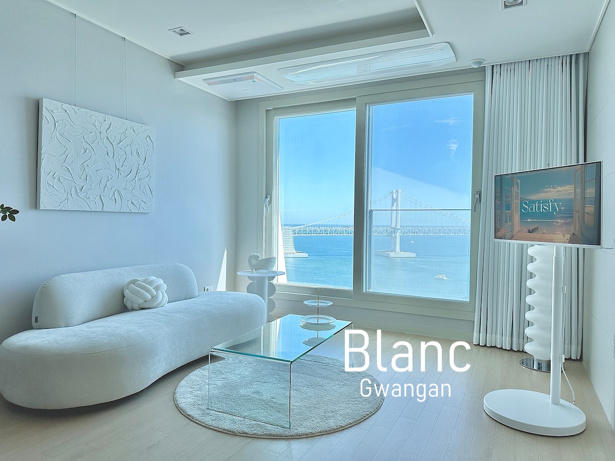 A bright living area features a plush white sofa and a glass coffee table, complemented by a scenic view of the Gwangan Bridge visible through large windows. A modern TV stand adds a sleek touch to the inviting space.