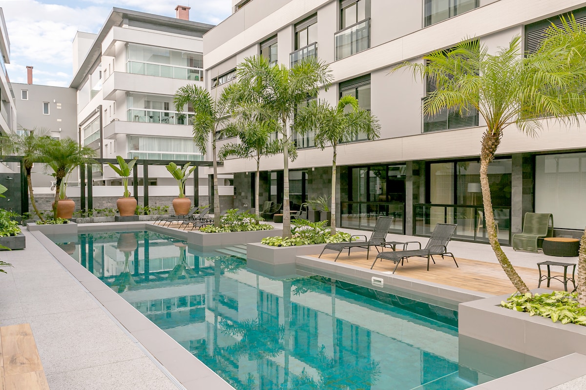 A modern outdoor pool area is surrounded by lush greenery and palm trees. Comfortable lounge chairs line the pool's edge, while planters add to the serene ambiance. The surrounding architecture features clean lines and large windows, enhancing the light and airy environment.
