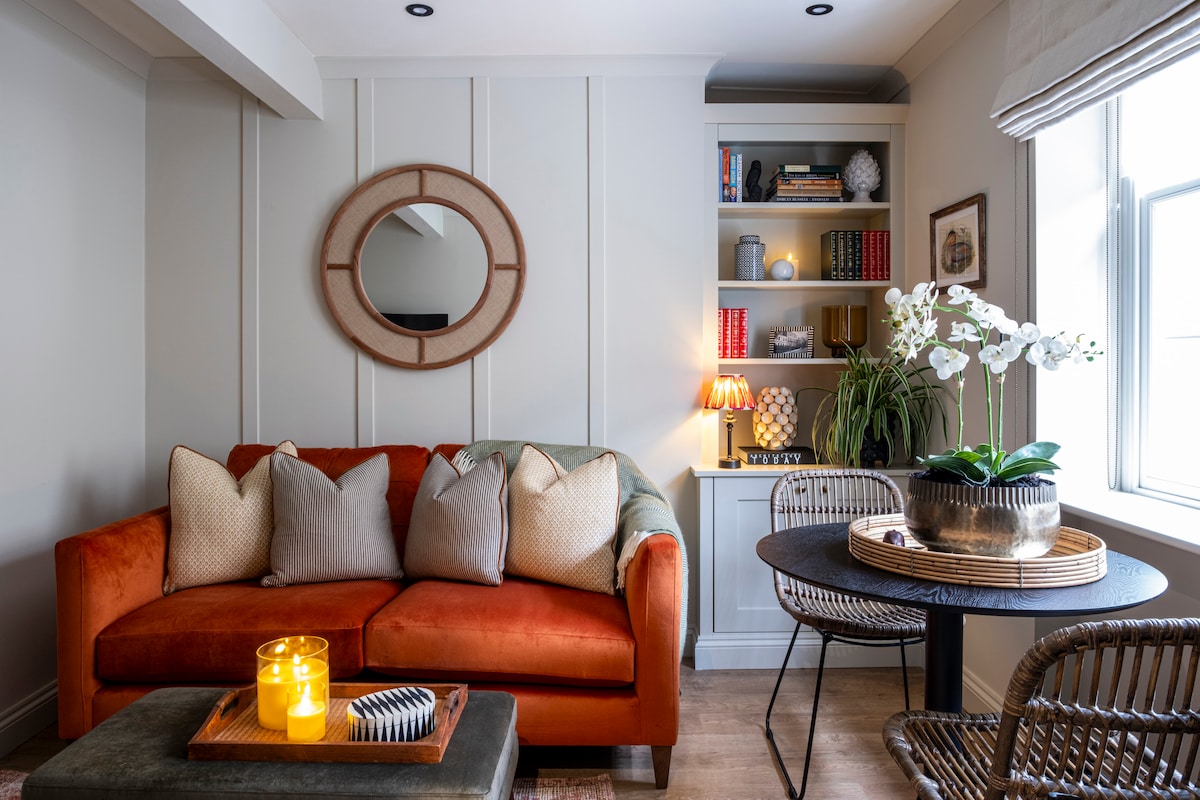A cozy living area is furnished with a vibrant orange sofa adorned with light-colored cushions. A round dining table with woven chairs is positioned nearby. A shelf filled with books and decorative items complements the space, highlighted by soft lighting and an arranged potted plant.