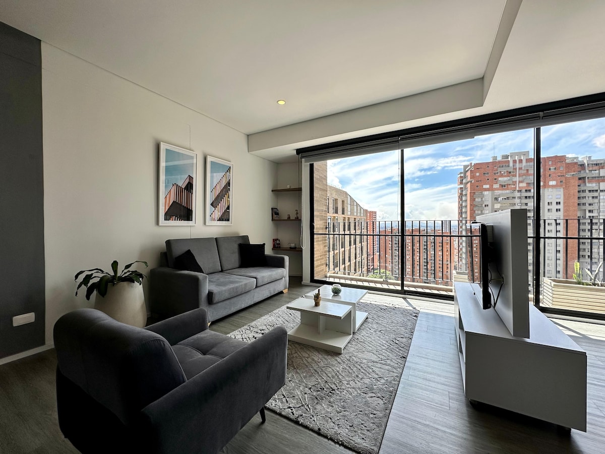 A modern living area is showcased with large windows providing expansive city views. A dark gray sofa and a matching armchair are positioned around a light coffee table. A textured area rug grounds the space, complementing a minimalist television stand and a decorative plant near the window.