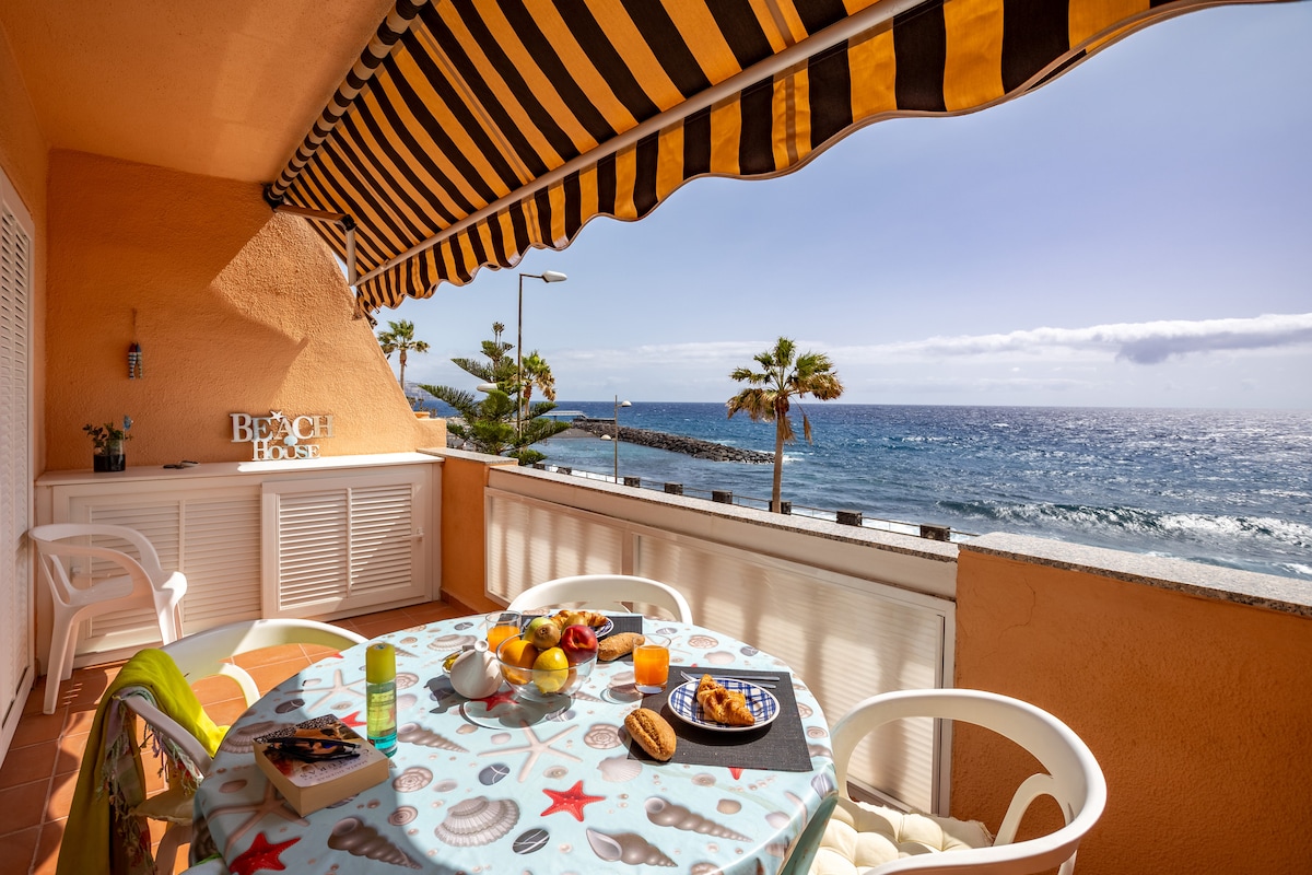 A private terrace features a round table set with various breakfast items, including fruits and pastries. White chairs surround the table, and a striped awning offers shade. The ocean is visible in the background, accompanied by palm trees, with sunlight illuminating the scene.