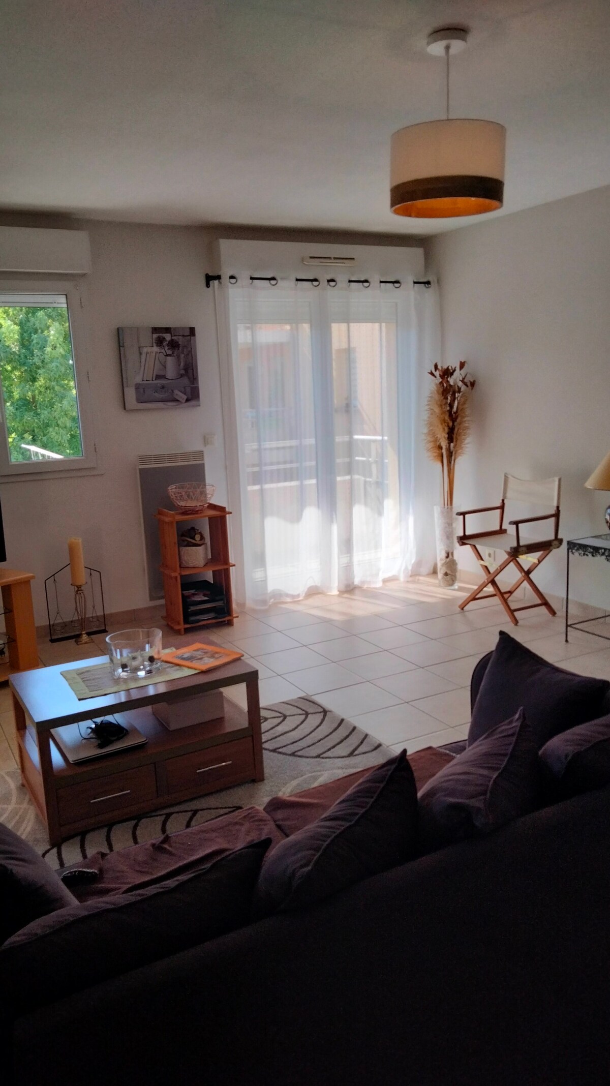 A bright living room is showcased with soft natural light entering through glass doors that lead to a balcony. A comfortable dark sofa is positioned alongside a wooden coffee table, and two chairs with a small side table are situated near the window.