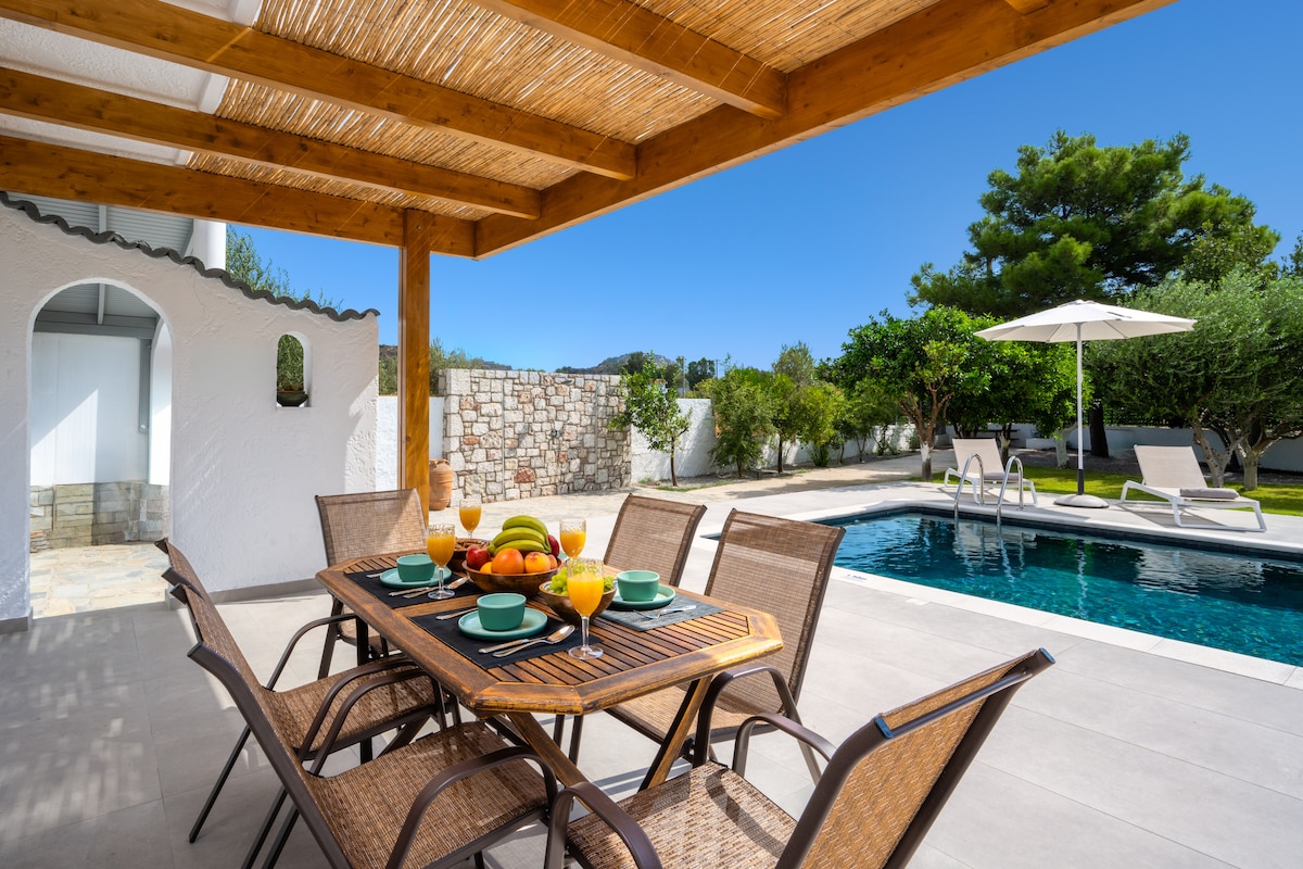 An outdoor dining area is presented under a wooden pergola, featuring a table set with colorful plates and fresh fruit. Nearby, a private swimming pool glistens under the sun, accompanied by comfortable loungers and an umbrella, all surrounded by lush greenery.
