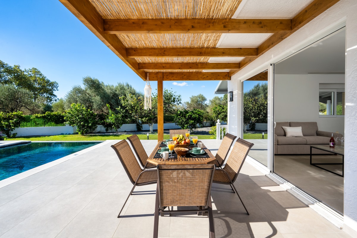 An outdoor dining area is visible under a wooden pergola, featuring a table set with colorful dishes and fresh fruit. Adjacent to the pool, the space overlooks a lush garden with well-maintained greenery, creating an inviting area for meals and relaxation.