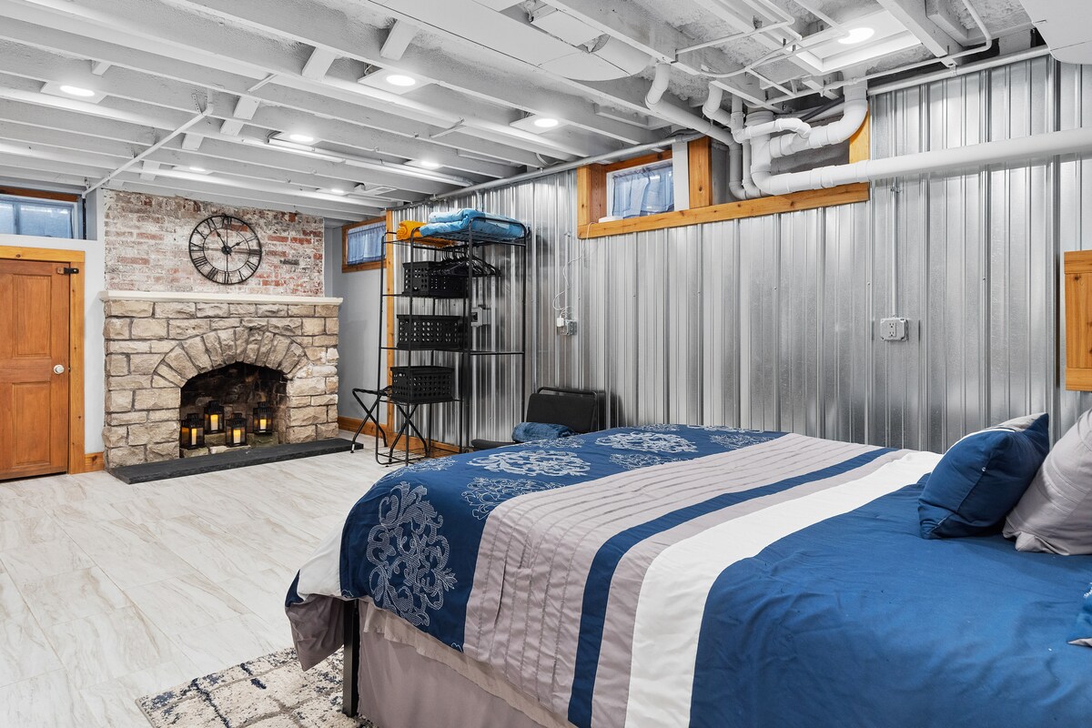 An industrial-style studio apartment features a queen-sized bed adorned with blue and grey bedding. A decorative stone fireplace is set against a backdrop of metal walls, with a clock and candle arrangement enhancing the space. Natural light enters through two small windows.
