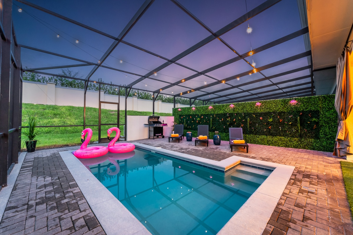 A private heated pool area is enclosed with a screen for safety. Two vibrant pink inflatable swan floats are positioned in the clear water. Relaxing lounge chairs are arranged nearby, accompanied by a barbecue grill and decorative greenery along the enclosure.