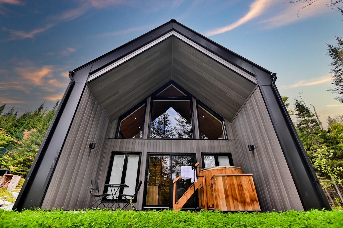 The exterior of a Scandinavian-style cottage is shown, featuring a unique angular design and large glass doors. A private cedar hot tub is placed on the deck, surrounded by lush green grass. Dense trees provide a natural backdrop, with a colorful sky at dusk highlighting the peaceful setting.
