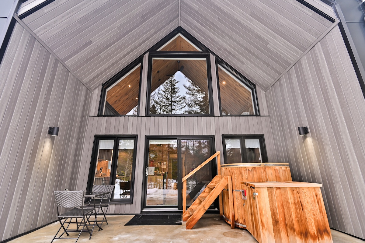 The entrance of the cottage features a modern design with high triangular ceilings and large windows offering views of the surrounding trees. A wooden staircase leads up to the main door, while two chairs are positioned on the patio for outdoor relaxation.