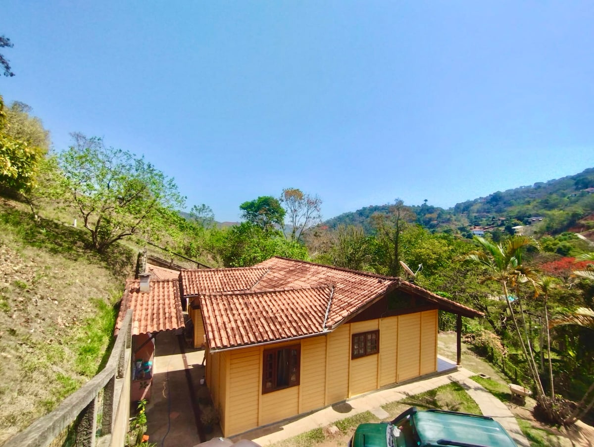 A quaint yellow house features a tiled roof and stands surrounded by lush greenery and hills. The outdoor area includes a pathway leading to the entrance, flanked by trees and shrubs, creating a serene and inviting environment.