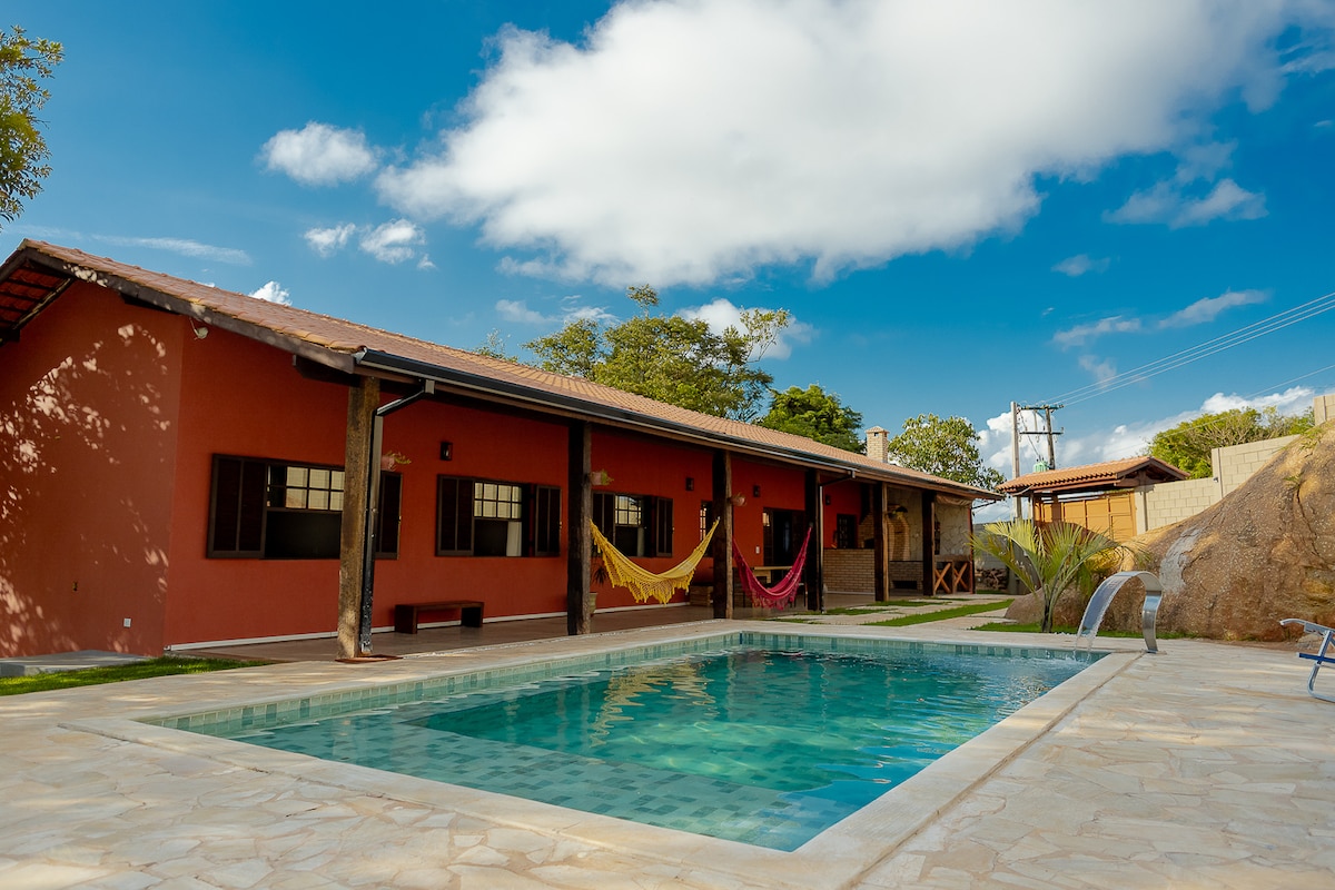 A spacious outdoor area features a bright pool with a cascading waterfall, surrounded by a stone patio. Hammocks are suspended from the porch overhang, inviting relaxation. The charming structure includes multiple windows, allowing ample natural light, and is set against a backdrop of lush greenery.