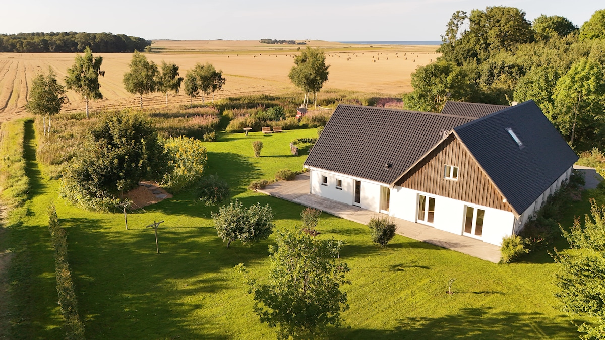 A modern villa is set amidst expansive green gardens, showcasing manicured landscaping. The property features a sleek roof and large windows, revealing the surrounding fields and distant coastline, creating a serene and inviting environment.