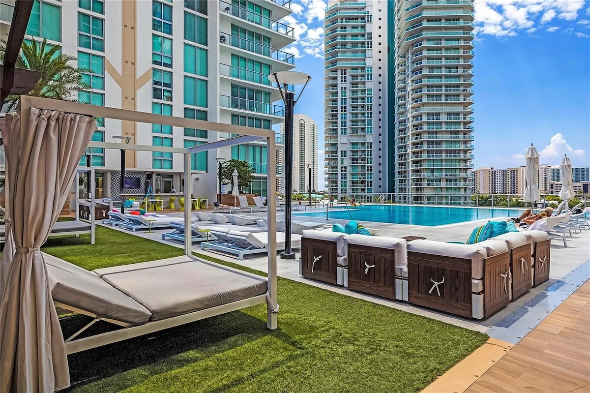 A sleek outdoor pool area is surrounded by modern high-rise buildings, featuring comfortable lounge chairs and daybeds. Lush green grass complements the space, along with shaded areas provided by umbrellas. The clear blue water of the expansive pool reflects the sunny sky.