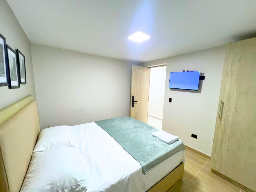 A bright and modern room features a spacious bed with a light green throw. A flat-screen television is mounted on the wall across from the bed. Soft lighting illuminates the space, while a wooden wardrobe stands nearby. A doorway leads to an adjoining area.