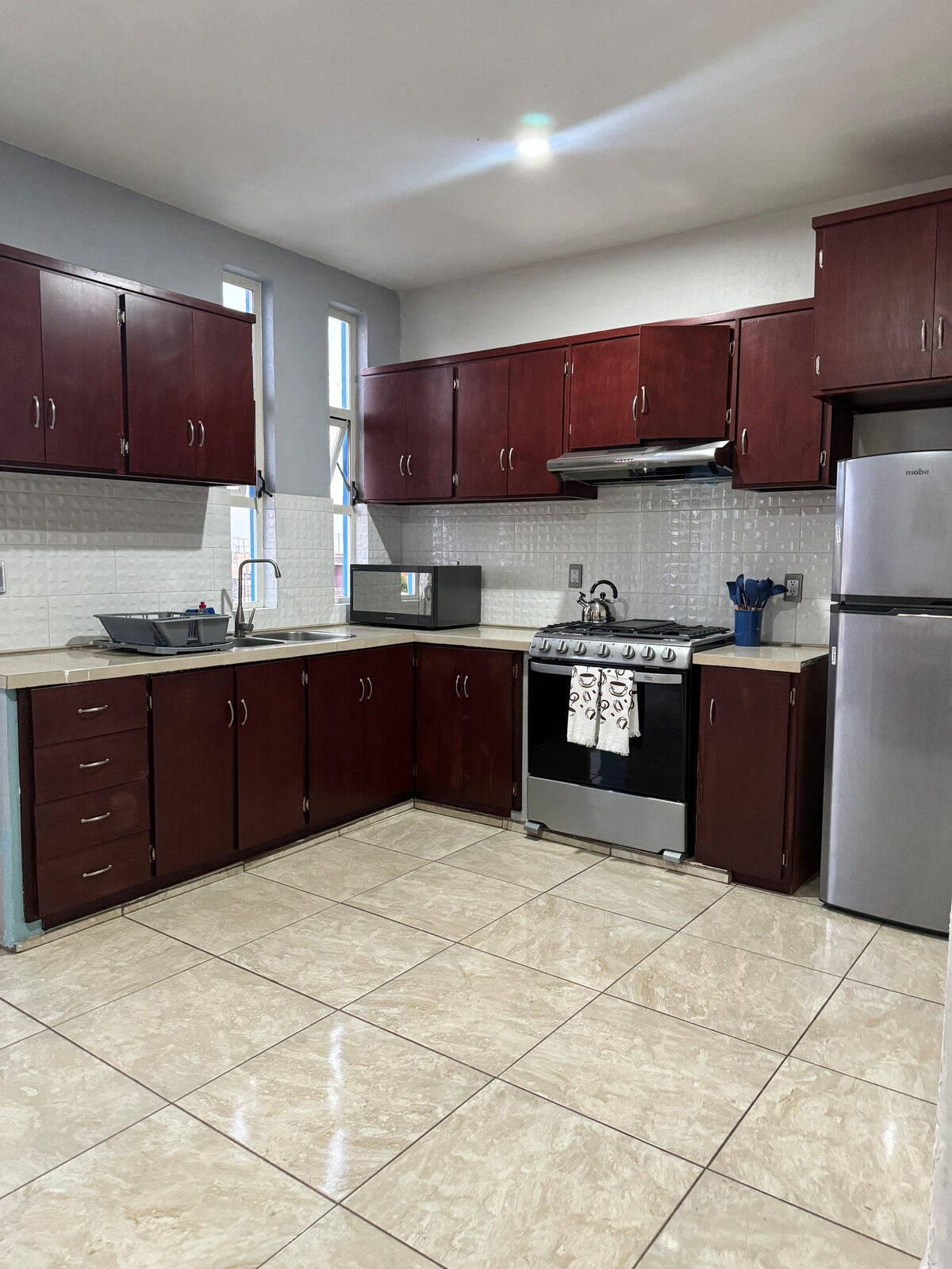A functional kitchen is depicted, featuring dark wood cabinetry and a modern stainless-steel stove. The countertop is equipped with a stainless-steel sink and a refrigerator, while tiles add a polished look to the floor. Natural light filters through the window, enhancing the workspace.