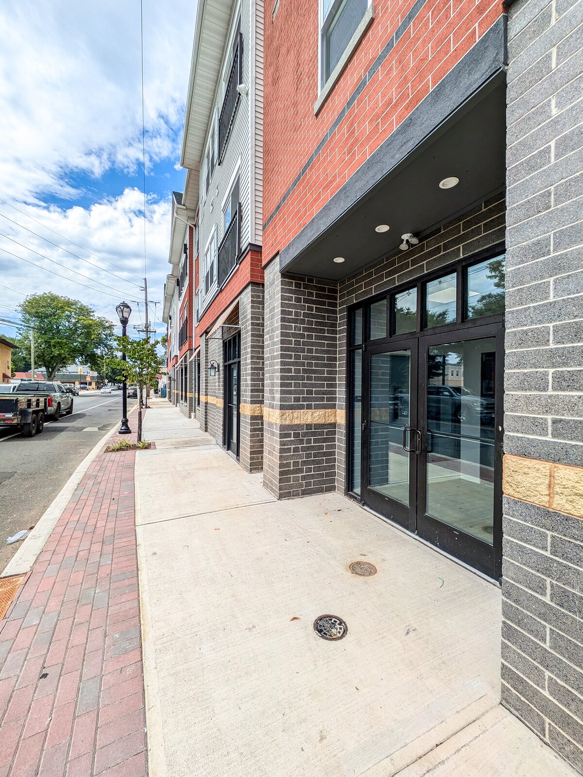 The image depicts the exterior of a modern building with a combination of red and gray brick finishes. Large glass doors are framed in black, leading to commercial spaces. A wide sidewalk is visible, bordered by decorative streetlamps and a well-maintained curb, typical of an urban area.