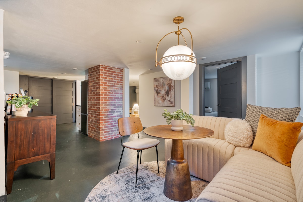 A modern living area features a curved sofa, a wooden table, and chairs. Textured throw pillows and a plush area rug enhance comfort. A brick column provides a rustic touch, while doors to the left lead to private rooms, ensuring a cozy and inviting space.