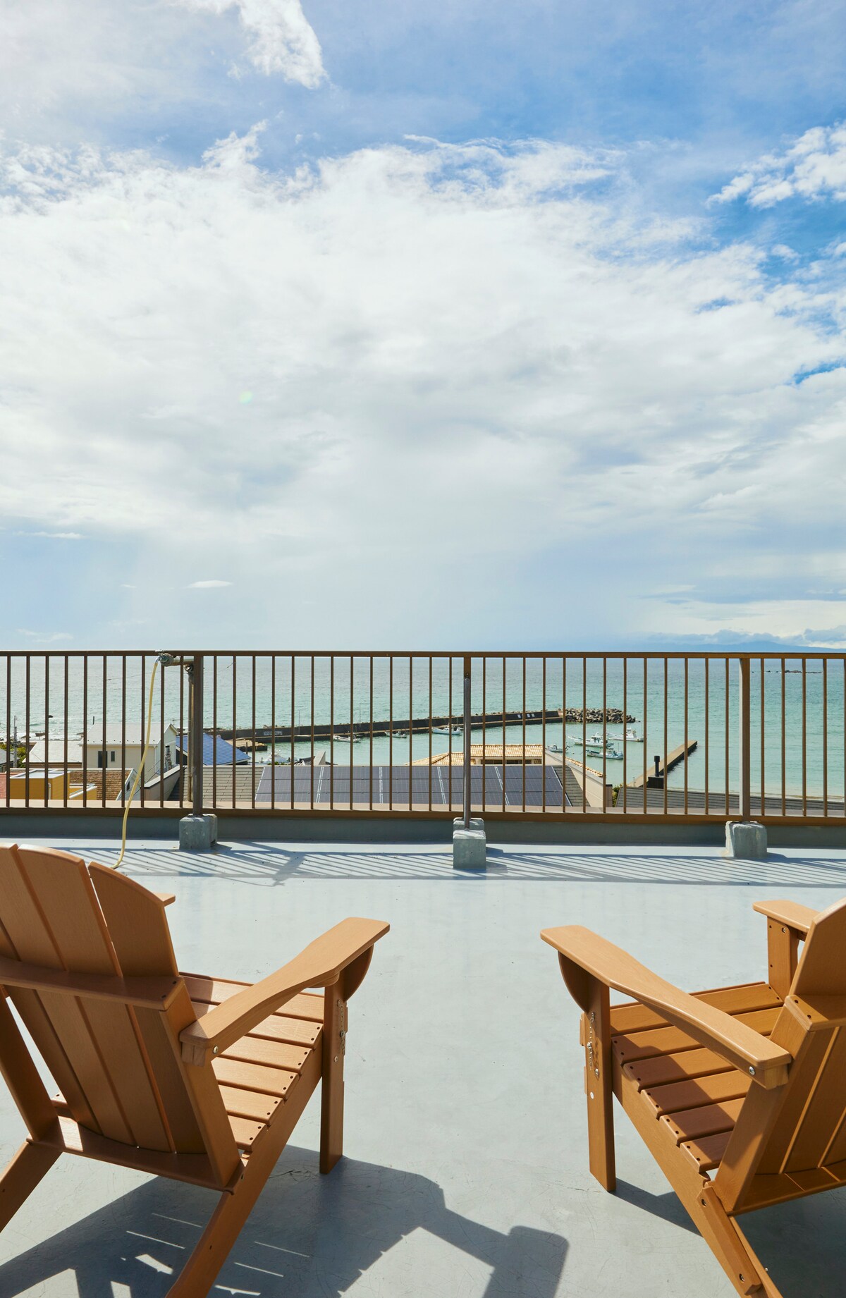 A spacious balcony is furnished with two wooden chairs, facing a scenic view of the ocean. The sky is filled with soft clouds, and sunlight reflects off the water. A railing provides safety while allowing unobstructed vistas of the serene landscape.