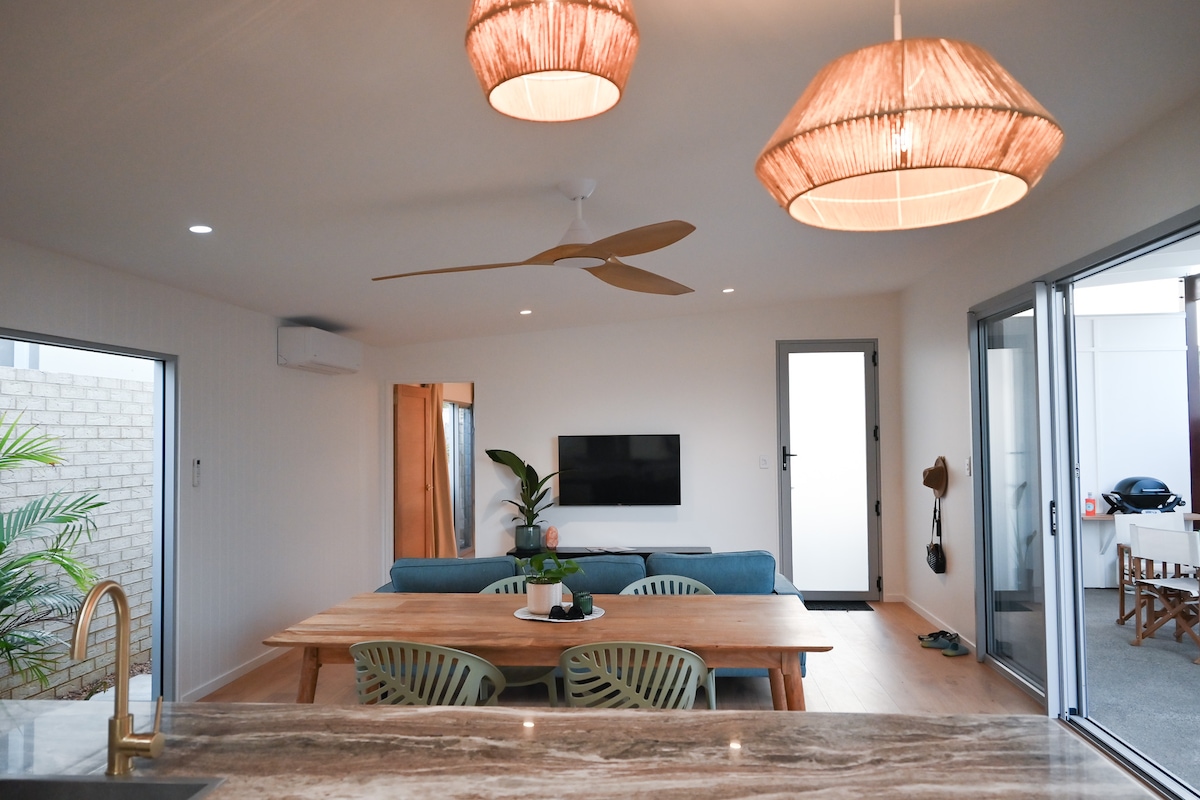 A contemporary open-plan living area is highlighted by a large wooden dining table surrounded by green chairs. A comfortable blue couch faces a wall-mounted television. Two woven pendant lights hang from the ceiling, and light pours in through sliding glass doors that connect to an outdoor space.