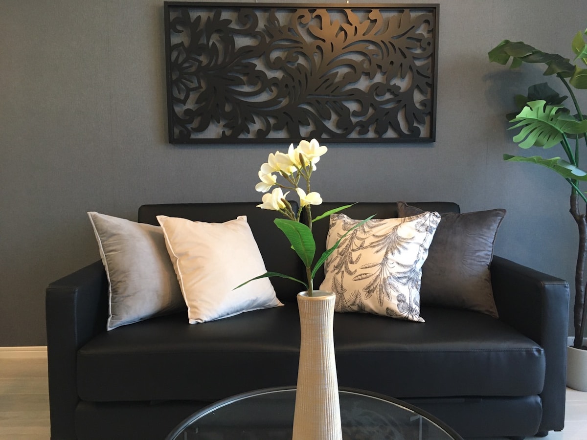 A black sofa is complemented by decorative pillows in muted tones. A tall vase with white flowers is placed on a glass coffee table in front of the sofa, which is set against a textured gray wall featuring an artistic panel.