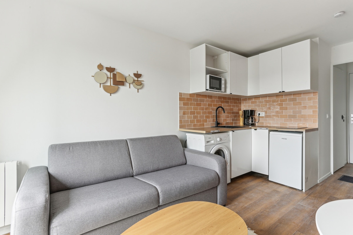 A welcoming living space is presented, featuring a gray sofa and a round wooden coffee table. A modern kitchenette with white cabinets includes a washing machine, microwave, and coffee maker, complemented by warm brown tiling that adds a subtle contrast.