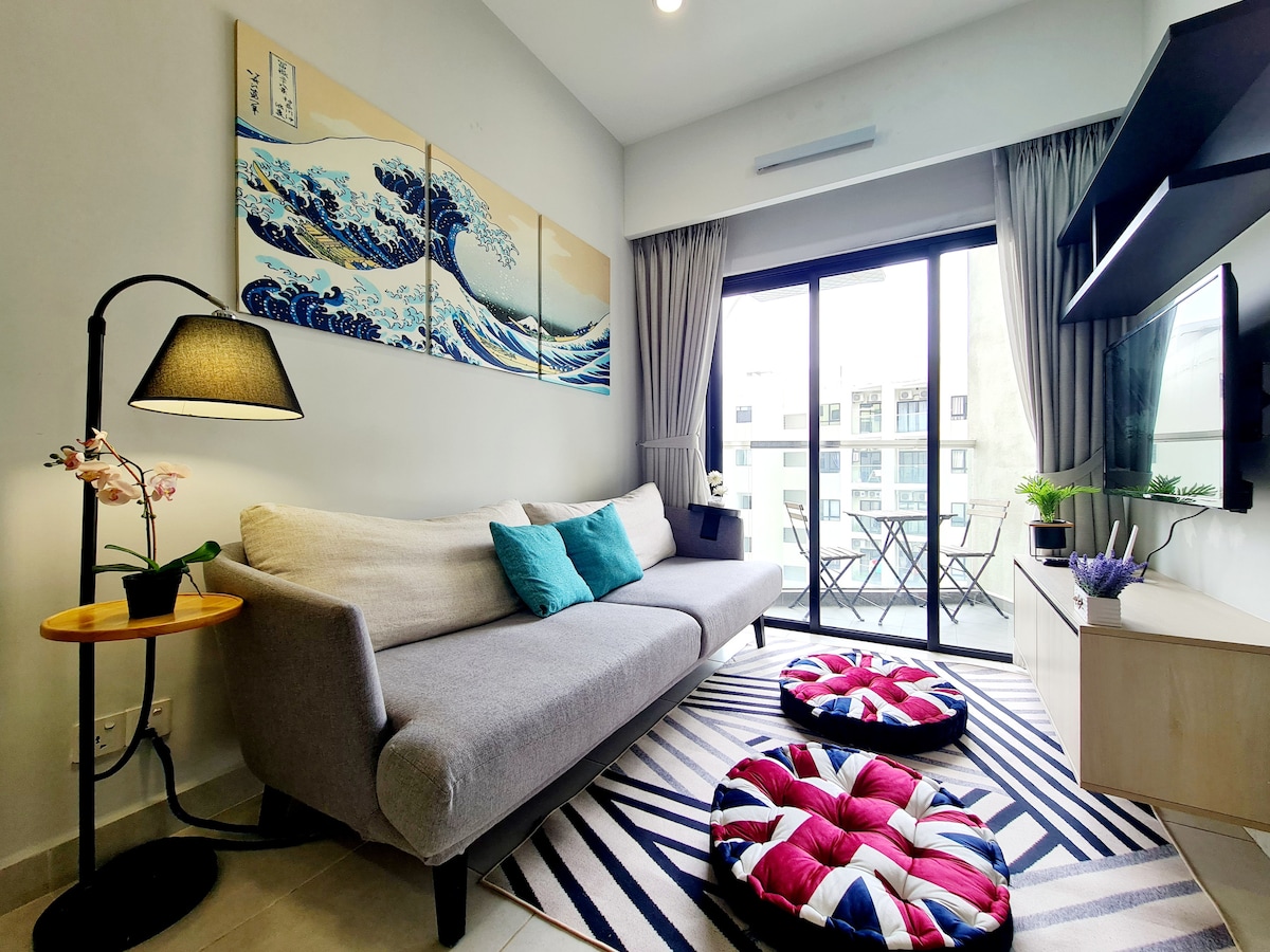 A comfortable living area is furnished with a gray sofa adorned with vibrant blue cushions. Floor-to-ceiling windows provide access to a small balcony with outdoor seating. A striped area rug features circular poufs, while an artwork depicting ocean waves decorates the wall.