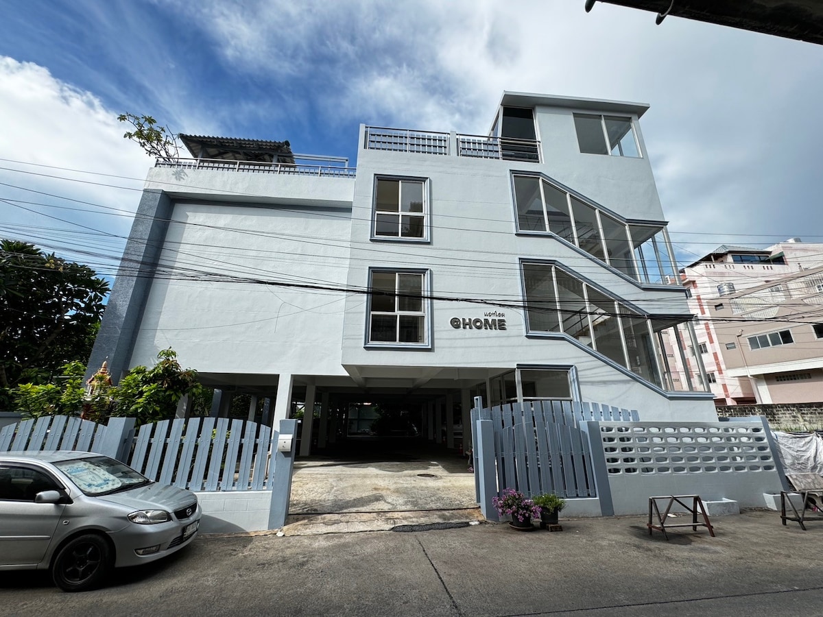 A modern, light blue building features multiple levels and large windows. An open parking area is located underneath, and a staircase leads up to the entrances. The surroundings include greenery and neighboring structures, contributing to a welcoming urban environment.