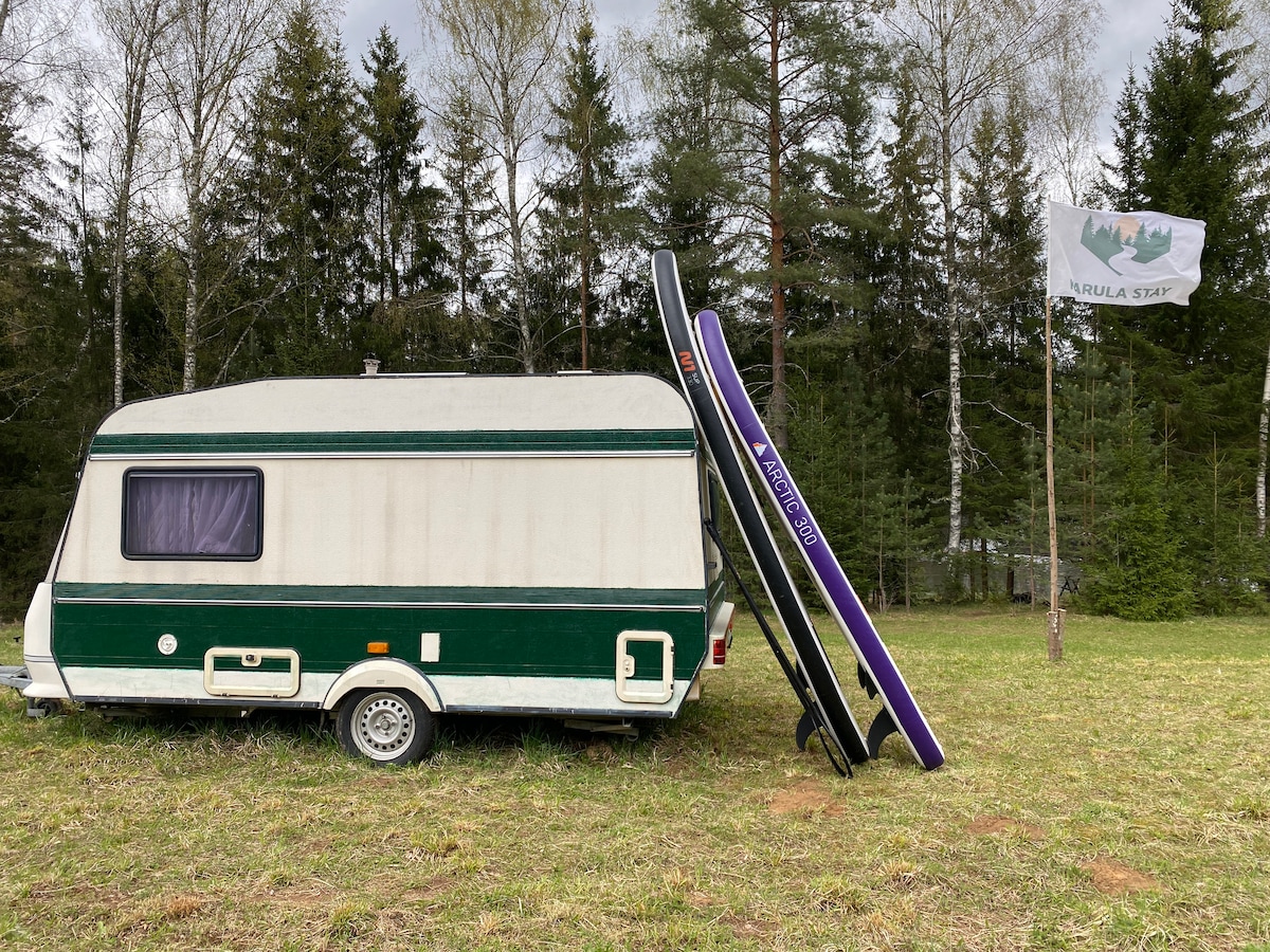A vintage camper is parked on a grassy area, surrounded by tall trees and greenery. Two stand-up paddleboards lean against the camper, and a flag indicating the location is visible. The overall setting conveys a natural, secluded environment.