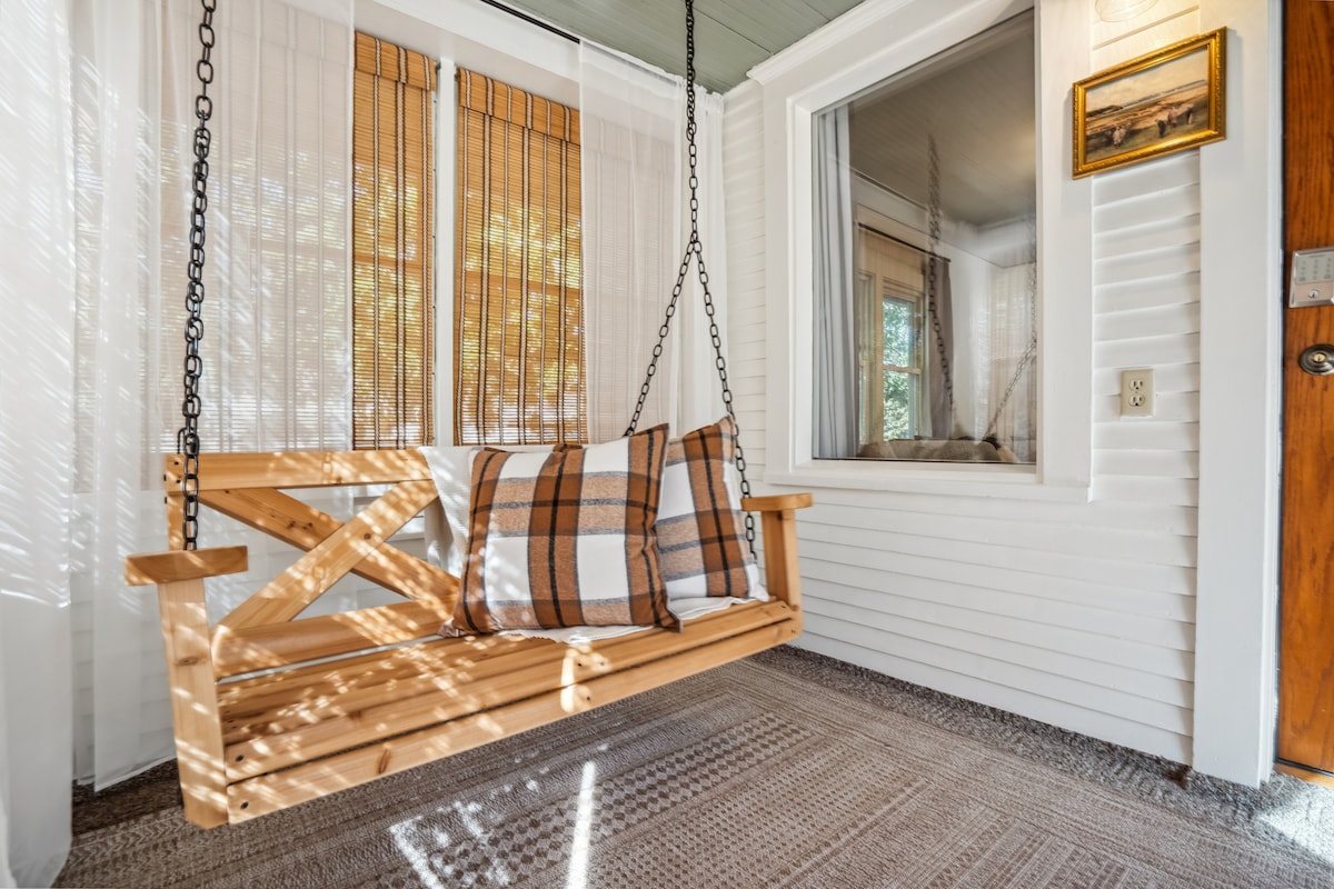 A wooden swing hangs from the ceiling in a sunlit porch area. Two pillows are placed on the swing, which is positioned against a light wall. Natural light filters through sheer curtains, casting soft shadows on the floor.