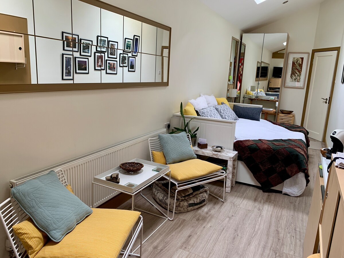 The interior of the studio presents a blend of functionality and comfort. A spacious bed is complemented by a cozy quilt and decorative pillows. Two lounge chairs with yellow cushions and a small table create a relaxing seating area. A mirror reflects the wall featuring framed artworks, enhancing the lively atmosphere.