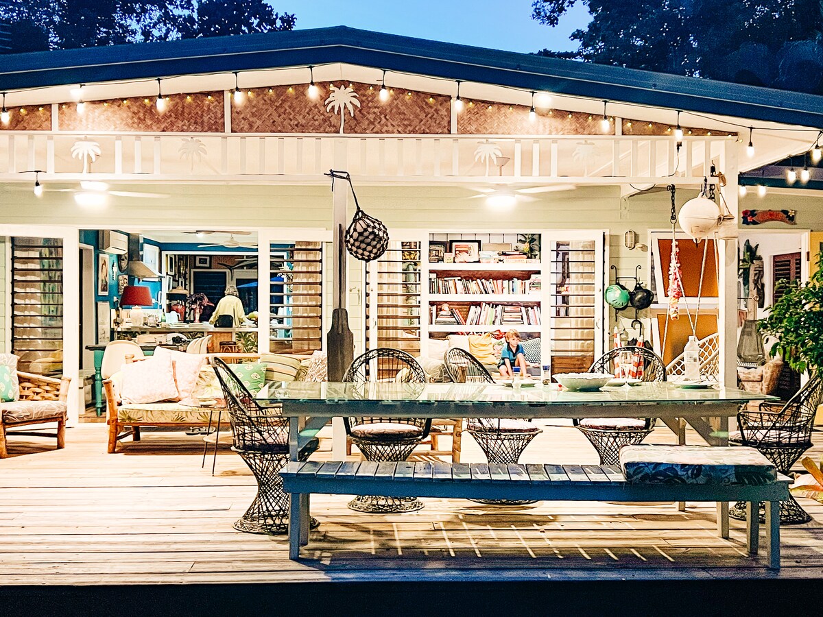 An outdoor dining area is illuminated by warm lights, featuring a long wooden table surrounded by woven chairs. The space opens up to a living area with eclectic decor, showcasing vibrant colors and ample seating. Tropical plants add natural greenery throughout.