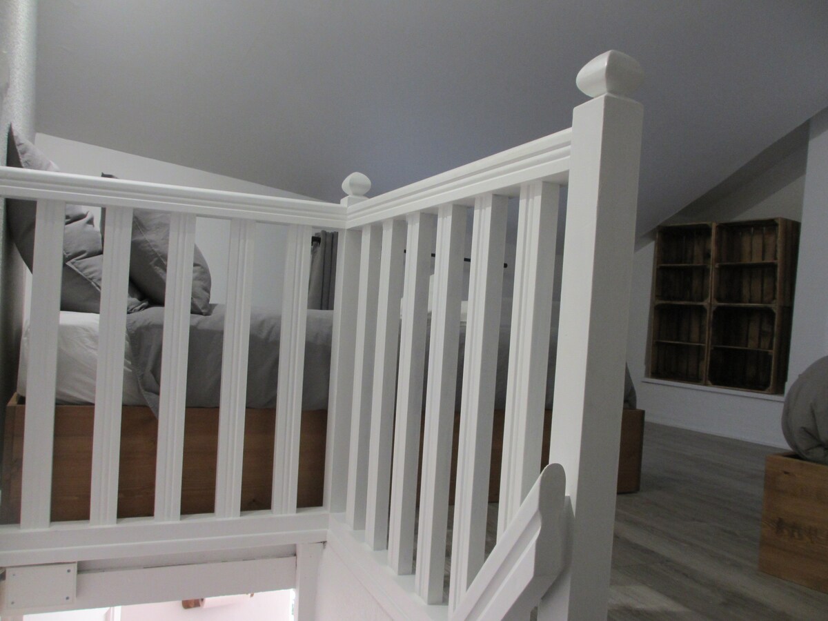 An upstairs area features a sturdy white railing leading to a sleeping space. A spacious bed with a neutral-toned comforter is visible at the top, complemented by gray pillows. Soft, natural light fills the room, enhancing the light wooden accents on the floor.