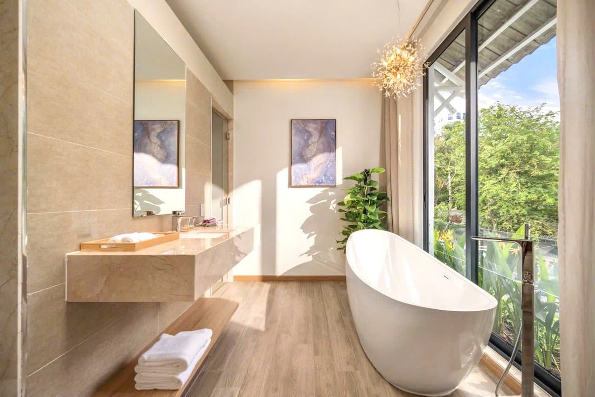 A modern bathroom features a freestanding soaking tub illuminated by natural light through expansive glass doors. A stylish vanity with dual mirrors complements the light wooden accents. Fresh greenery is seen outside, enhancing a serene atmosphere.