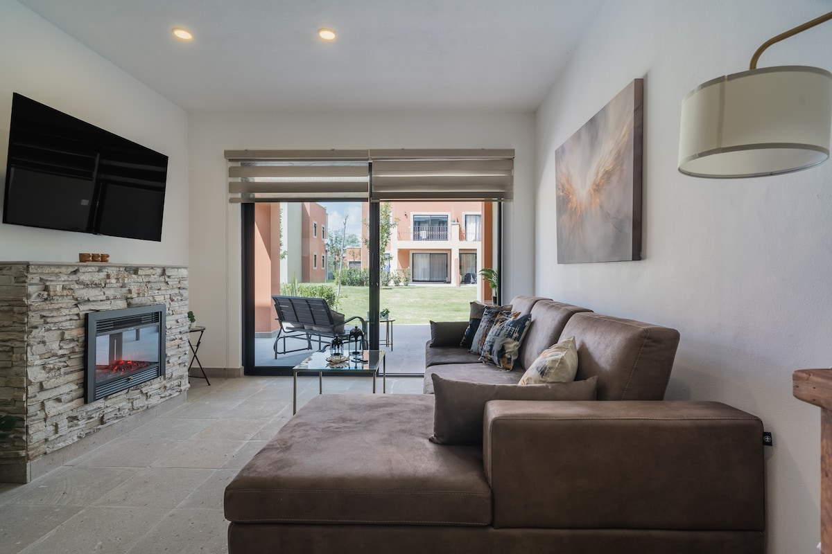 A comfortable living area features a spacious brown sectional sofa and a glass coffee table positioned in front of a modern electric fireplace. Large glass sliding doors lead to a view of the outdoor space, while soft lighting and neutral tones create a relaxed environment.