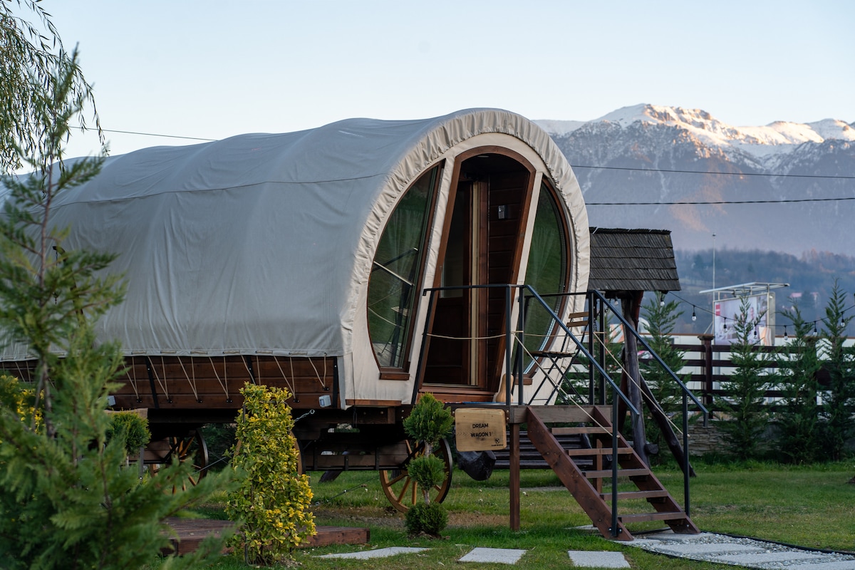 A rustic wooden caravan is showcased, featuring a curved canvas top and large front windows. Steps lead up to the entrance, inviting visitors to explore. Surrounding greenery adds to the natural setting, with distant mountains visible in the background under a clear sky.