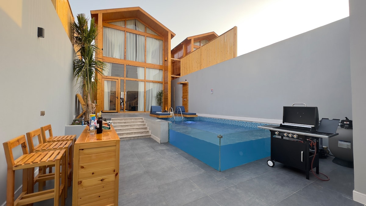 An outdoor area featuring a crystal-clear pool surrounded by a stone patio. A wooden bar set with stools is positioned nearby, and a barbecue grill is visible. Tall plants provide a touch of greenery beside the pool, enhancing the inviting ambiance.
