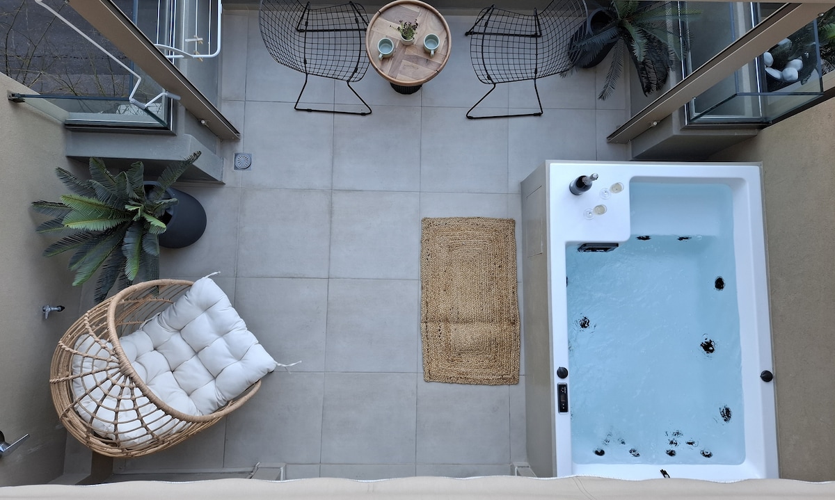 An outdoor balcony highlights a private heated jacuzzi surrounded by modern decor. Nearby, a comfortable hanging chair is positioned next to a small table with two chairs. Green plants add a natural touch to the relaxing space.