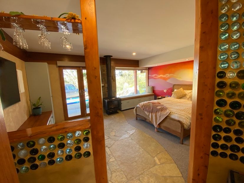 An open-plan bedroom is presented, featuring a queen bed with a sunset mural behind it. Natural light filters through large windows, reflecting off the unique bottle walls. A wood heater is located nearby, and a TV is mounted on the wall facing the bed.