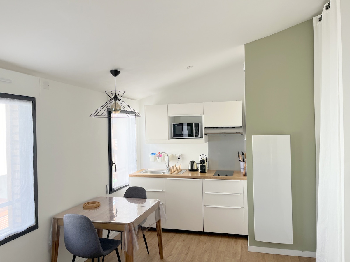 A modern kitchen area is depicted, featuring sleek white cabinetry and a built-in microwave. A wooden dining table with two gray chairs sits nearby. Natural light enters through a large window, with sheer curtains softly diffusing the sunlight.