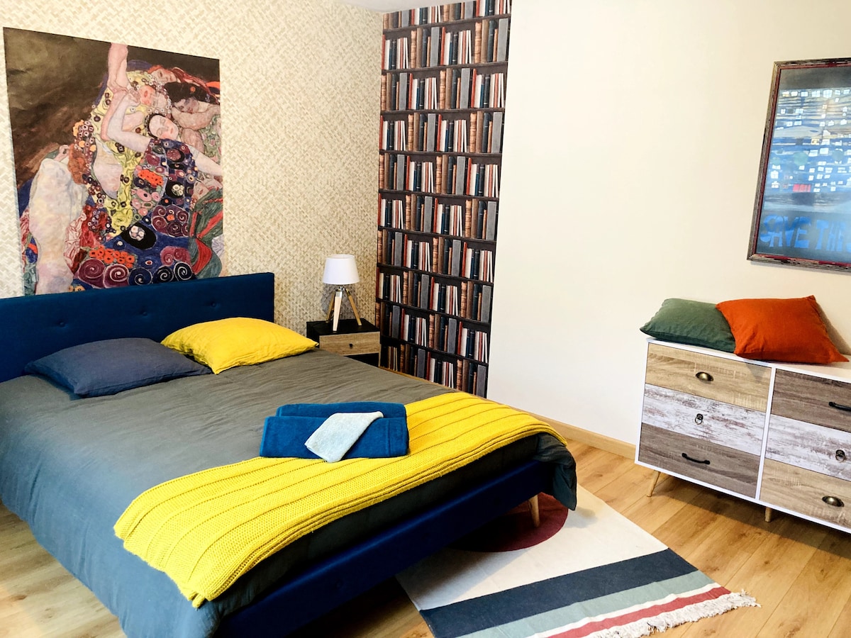 A spacious bedroom features a blue bed with a colorful yellow and grey blanket. An art piece adorns one wall while a bookshelf wallpaper adds texture. A bedside lamp stands next to a wooden nightstand, and a dresser with vibrant cushions completes the cozy atmosphere.