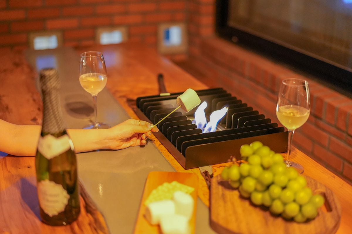 A hand is shown holding a skewer with a marshmallow over a small, stylish flame. Two glasses of wine accompany a bottle of sparkling wine and a wooden tray filled with grapes and other snacks. The setting is comfortably warm with a brick wall in the background.